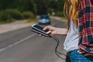 Nuori nainen seisoo tiellä pitäen kädessään kahta älypuhelinta, joista toinen on liitetty latauslaitteeseen. Taustalla näkyy auto ja vihreitä puita.