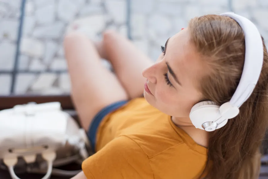 Nainen istuu rentoutumassa, päällään vaaleat vastamelukuulokkeet, kuunnellen musiikkia keltaisessa t-paidassa.